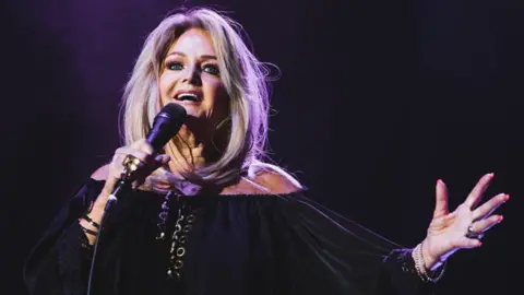 Getty Images Bonnie Tyler on stage singing. She wears a black long sleeve dress and holds the microphone in one hand, with the other outstretched. Her long blonde hair is loose. 