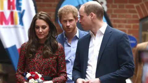 Reuters The Princess of Wales, dressed in a red floral dress, and Prince of Wales, in a blue suit, stand in front of the Duke of Sussex