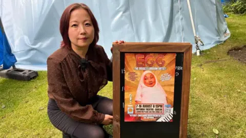 Sachie Mikawa A person crouches on a grassy area beside a wooden sandwich-board sign displaying a colorful poster for EGG: The Biggest Unborn Artist. The person is wearing a brown long-sleeved blouse with a bow at the neck.