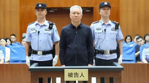 Shenzhen Intermediate People’s Court/WeChat China Evergrande Group founder Hui Ka Yan takes the stand in a court in China. He has a blank look and is standing between two officers, with members of the audience in the background.