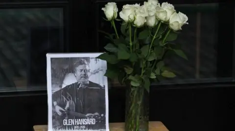 Damien Eagers/PA Wire A black and white picture of Glen Hansard sits on a table with a vase of white roses beside it