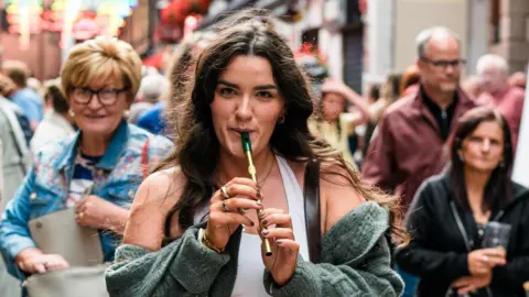 Belfast City Council A woman with long brown hair, playing a tin whistle. There is a crowd of people behind her. 
