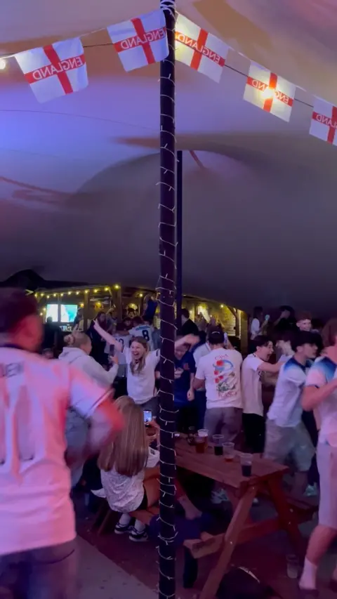 A group of fans sit on picnic tables underneath a white marquee with bunting of the England St George's cross. The fans are cheering in celebration at a goal. They are all wearing white England football shirts. 