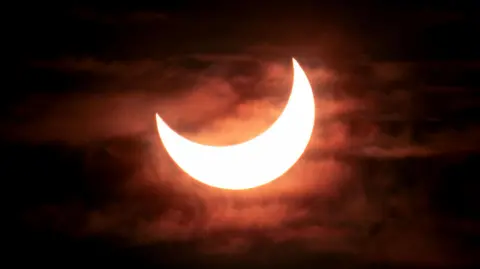 AFP via Getty Images A partial solar eclipse showing the bright crescent of the sun as the moon moves across it from the top left corner. The sky is dark with clouds visible behind the sun