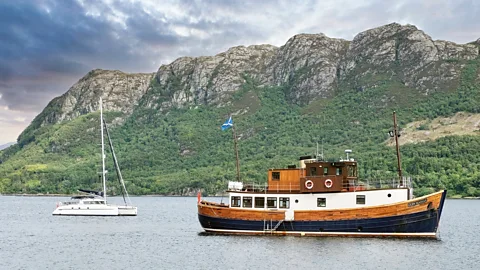 Alamy A small cruise boat and a yaught in front of a craggy and green hill (Credit: Alamy)
